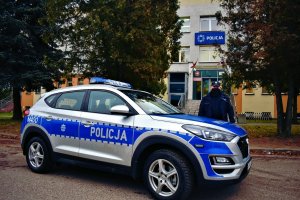 Radiowóz. Obok stoi policjant. W tle Komenda Powiatowa Policji w Augustowie.