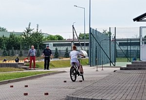 policjant stoi i tłumaczy dzieciom przepisy ruchu drogowego. chłopiec zdający egzamin na kartę rowerową jedzie slalomem, chłopiec zdający egzamin na kartę rowerową jedzie rowerem między pachołkami