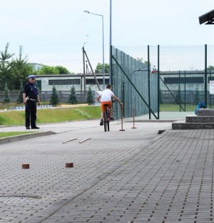 policjant stoi i tłumaczy dzieciom przepisy ruchu drogowego. chłopiec zdający egzamin na kartę rowerową jedzie slalomem, chłopiec zdający egzamin na kartę rowerową jedzie rowerem między pachołkami